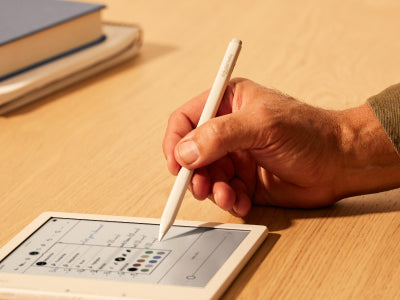 A hand is shown holding a Kobo Stylus 2 poised over a Kobo Libra Colour eReader on a flat surface. The tip of the stylus is about to select one of a range of pen colours shown in an options menu on the eReader screen.