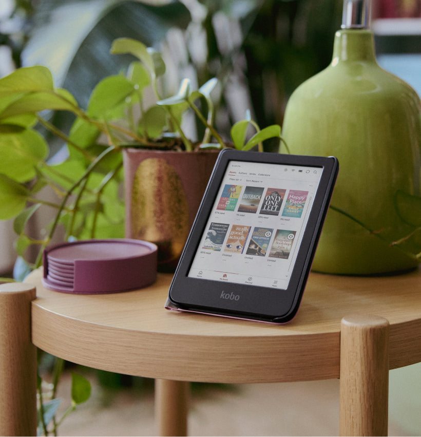 A Kobo Clara Colour eReader in its SleepCover stand, set on a wooden table by a potted plant and lamp.