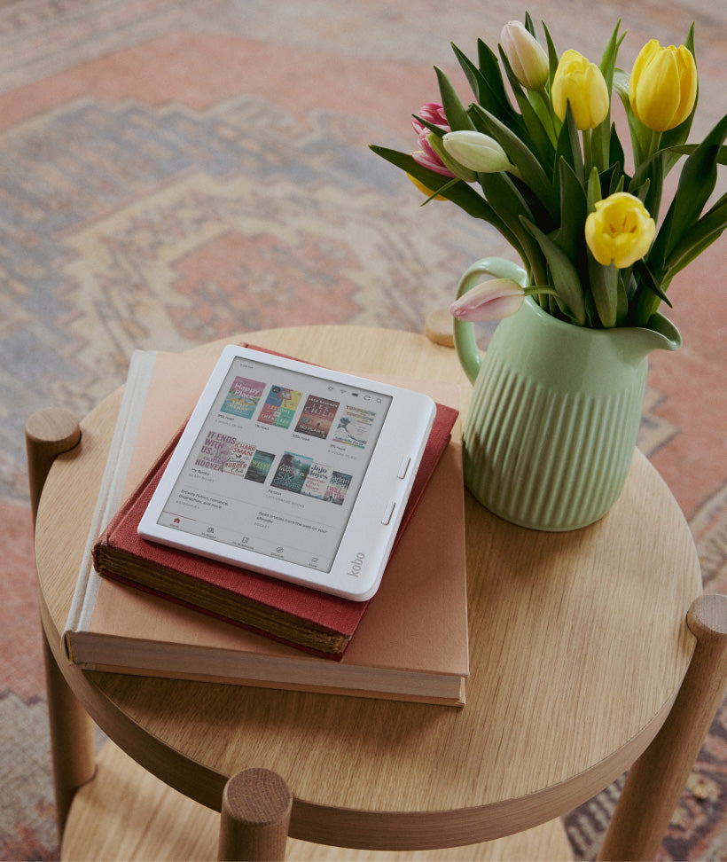 A Kobo Libra Colour eReader sitting on two hardcover books, set on a wooden table by flowers in a green vase.