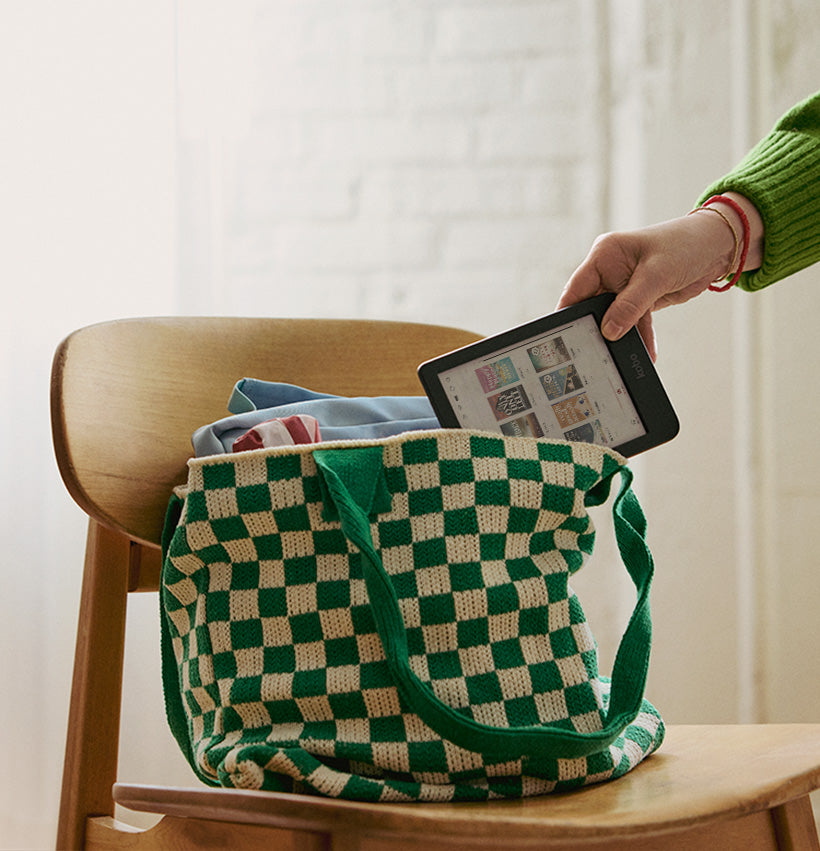 A hand removes a Kobo Clara Colour eReader from a bookbag sitting on a wooden chair.